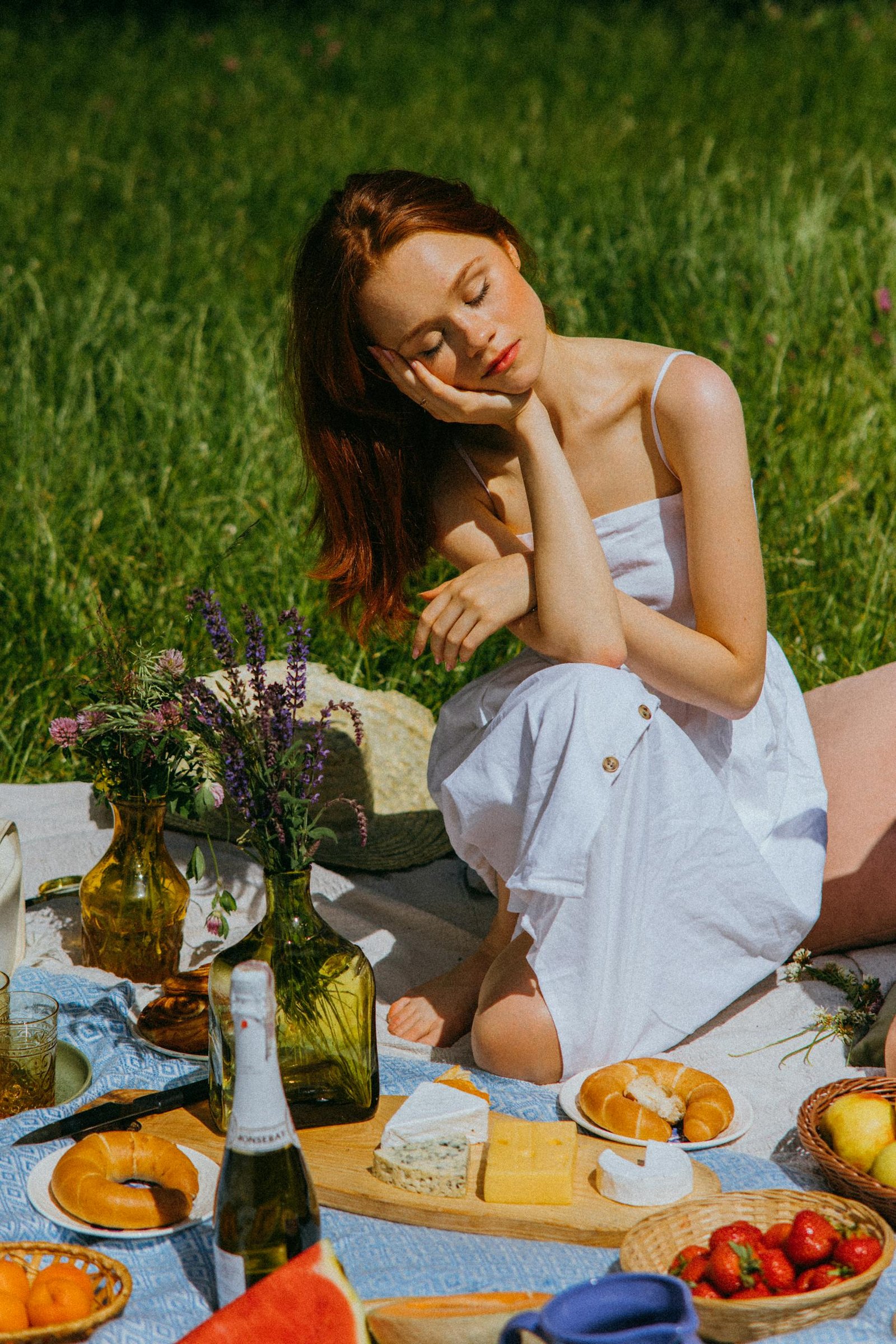 A tranquil summer picnic scene featuring a woman in a white dress, surrounded by delicious foods and wine.